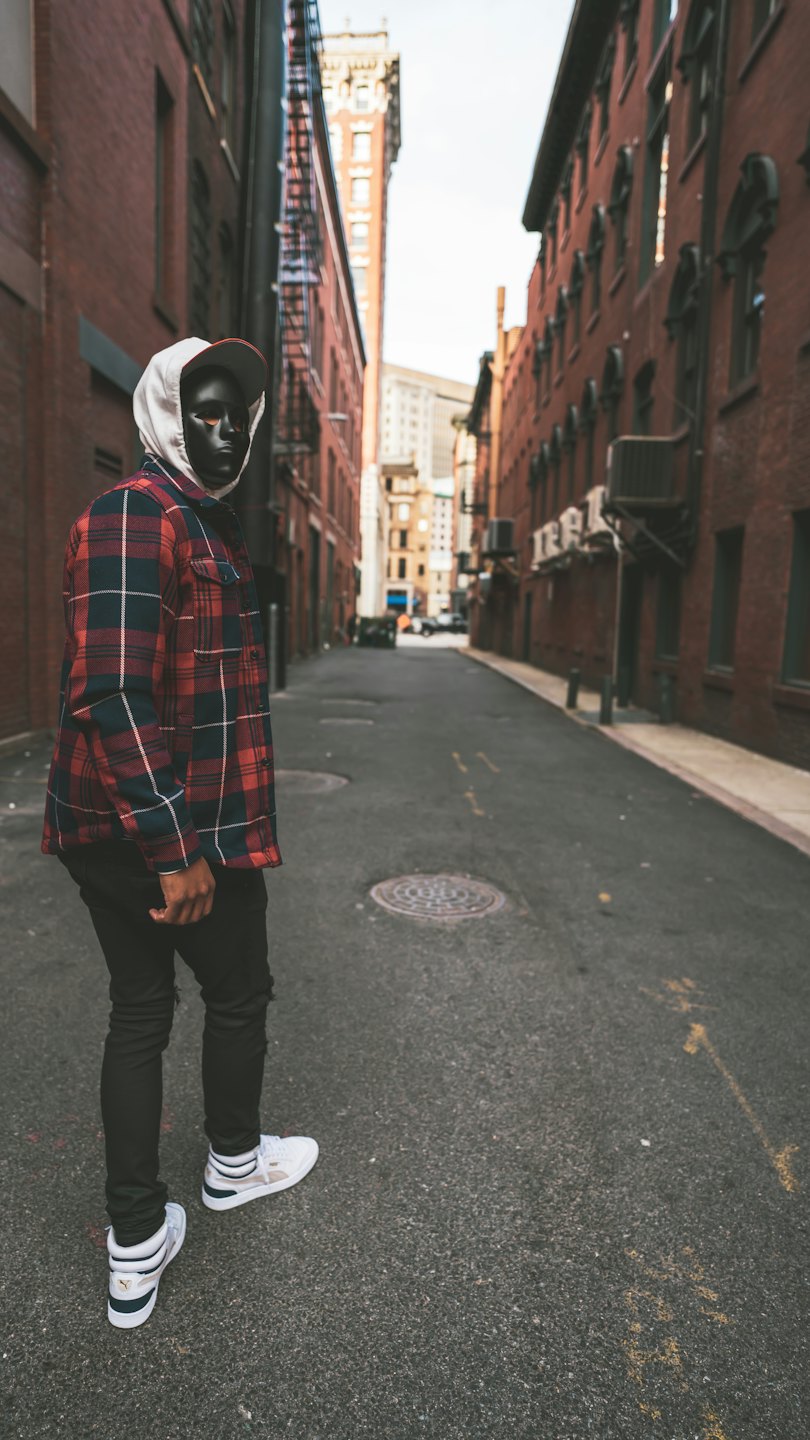 Person in plaid jacket and mask stands in urban alleyway.