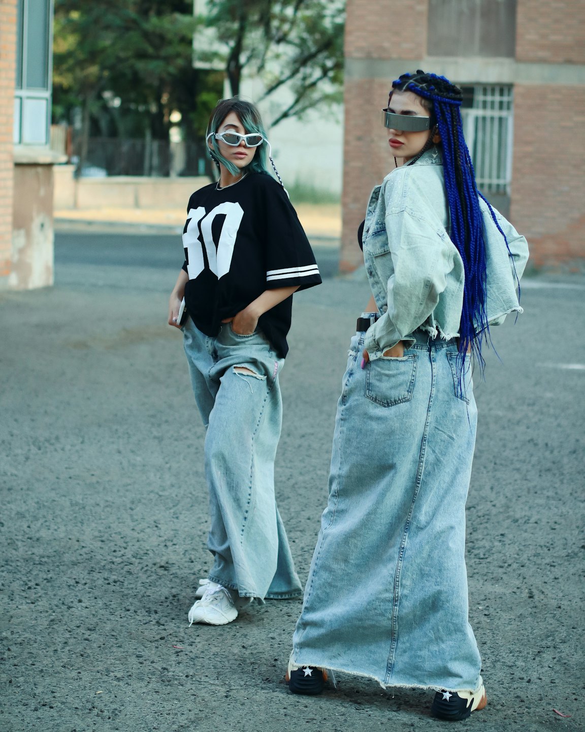 Two women in trendy streetwear pose on a paved lot.