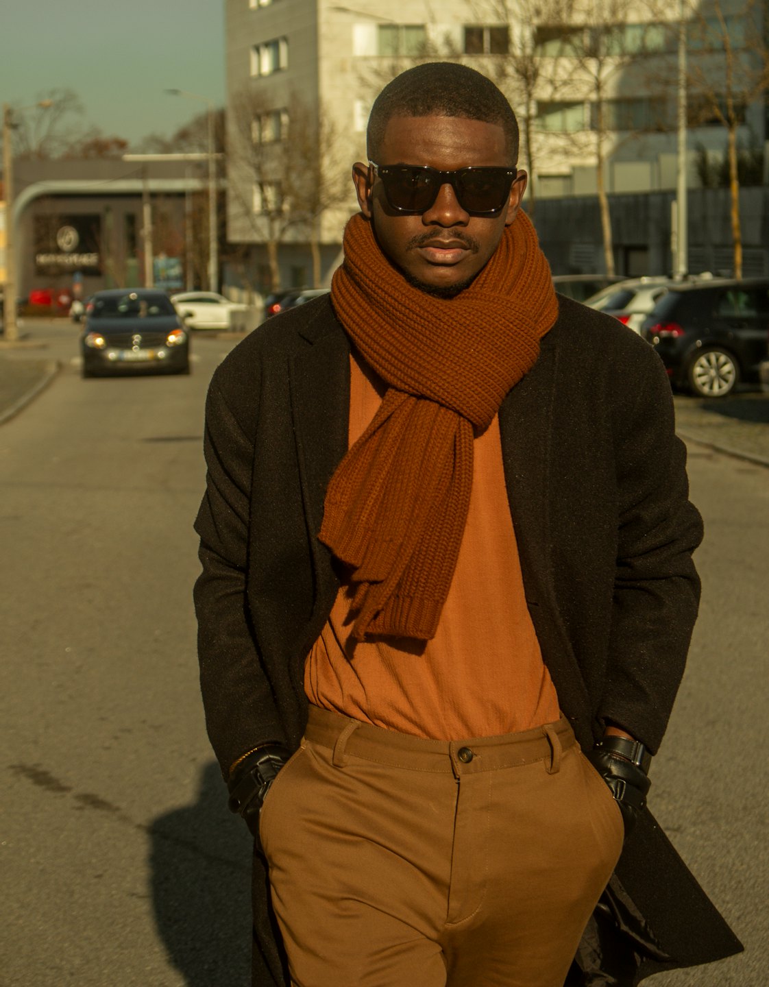 Man in stylish coat and scarf stands outdoors on city street.