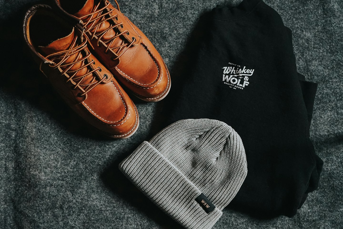 Brown leather moc-toe shoes, black sweatshirt, and grey beanie arranged on fabric.